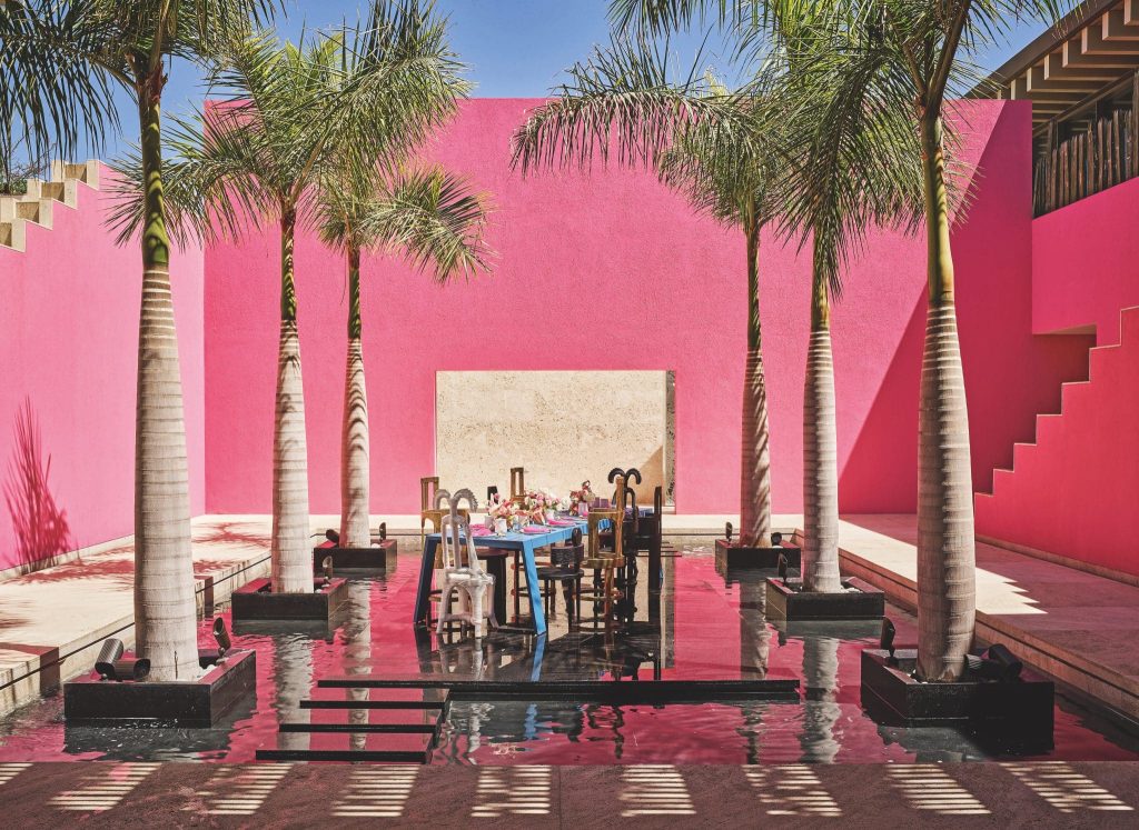 The Baja House, a pink building with palm trees and a table centered in a pool of water
